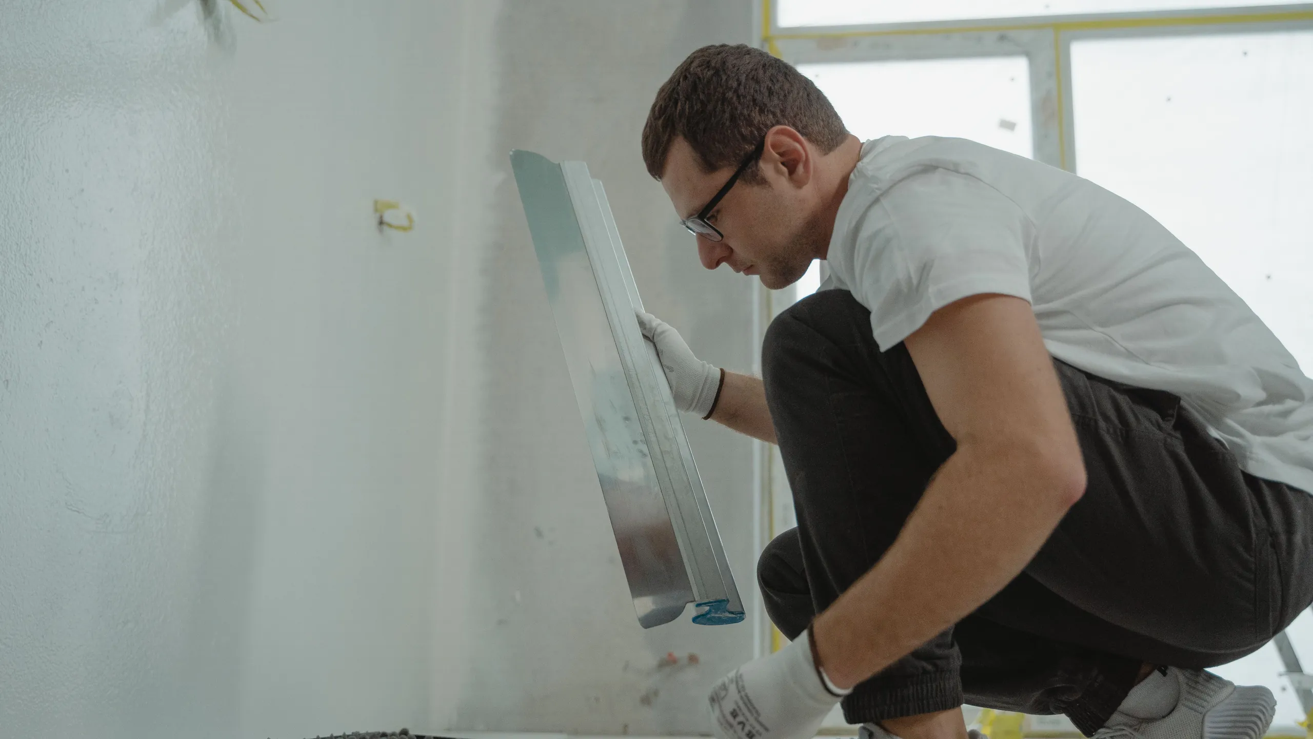 Handwerker mit Arbeitshandschuhen zieht eine lange Glättkelle über eine weiß gespachtelte Innenwand, Werkzeugkoffer am Boden.